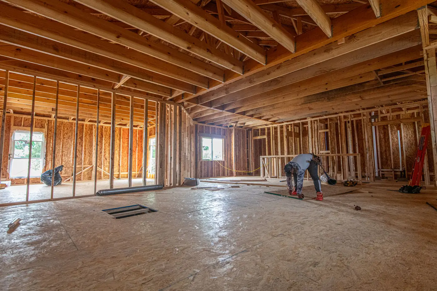Worker inside a wood-framed house with open stud walls, floor joists and a plywood subfloor