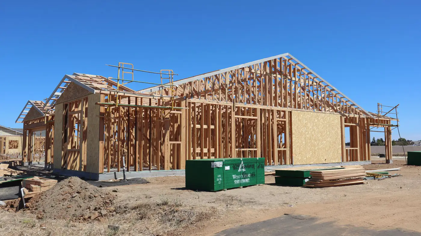 Single-story house framed in wood with stacked lumber and a pile of dirt on the lot under a clear sky