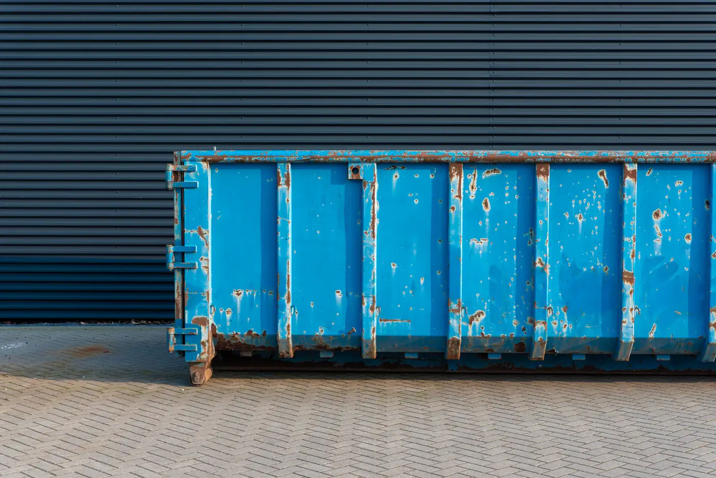 Weathered blue open-top roll off container parked on brick pavers beside a metal building