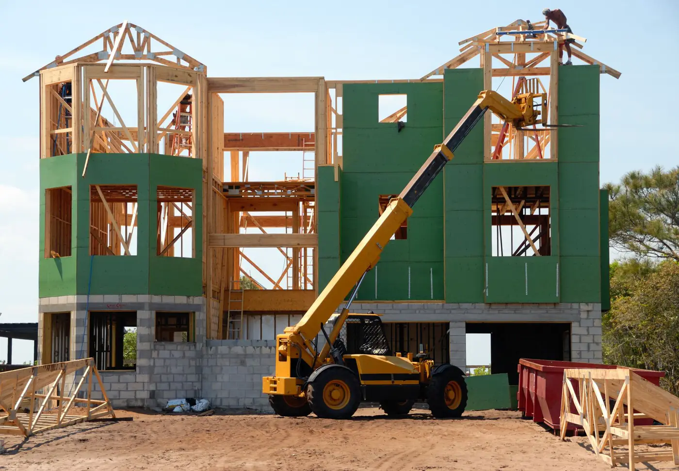 Red roll off container beside a wood-framed house under construction with a telehandler lifting materials