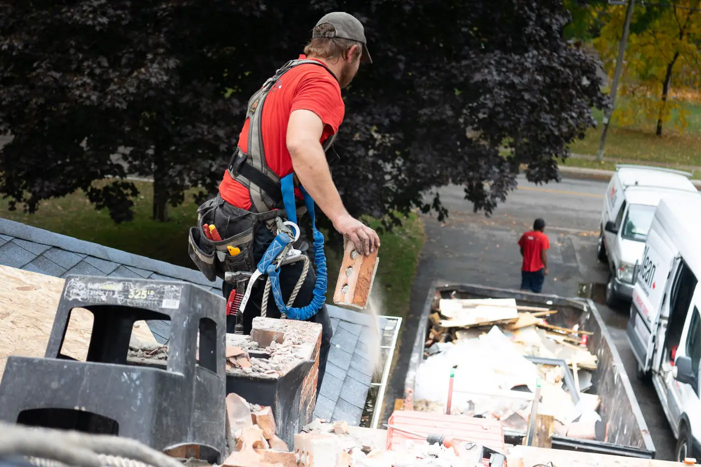 Roofer in a safety harness tossing old brick from a chimney into a container filled with roofing debris in the driveway below