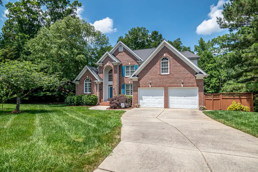 Brick two-story home with a two-car garage and a long concrete driveway surrounded by tall trees