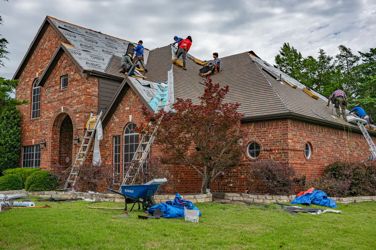 Roofing crew replacing the roof on a large brick house, with ladders, tarps and a wheelbarrow on the lawn